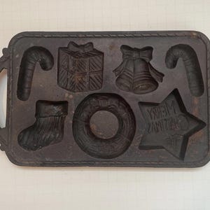 May include: A cast iron Christmas-themed baking mold with handles. The mold features shapes of a candy cane, gift box, bell, stocking, wreath, and a star with the text "MERRY CHRISTMAS".