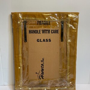 May include: A rectangular framed item, wrapped in clear plastic, with the text "FRAGILE HANDLE WITH CARE GLASS". The frame is a light brown colour, and the cardboard backing has the logo "Homco, Inc."