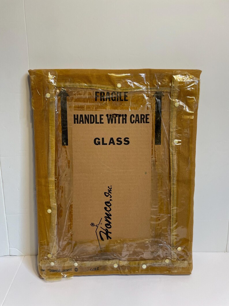 May include: A rectangular framed item, wrapped in clear plastic, with the text "FRAGILE HANDLE WITH CARE GLASS". The frame is a light brown colour, and the cardboard backing has the logo "Homco, Inc."