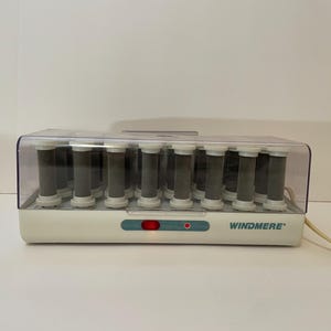 Op de afbeelding: Een witte en heldere Windmere haarroller set met 18 rollers. De basis heeft een turquoise accent met een rood licht, en de merknaam staat erop. De rollers zijn grijs en wit.