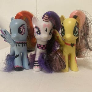 Könnte beinhalten: Drei bunte Spielzeugponys mit langem, gestyltem Haar. Die Ponys sind hellblau, weiß und gelb, jedes mit einzigartigen Markierungen und Accessoires. Das blaue Pony hat Flügel, während das gelbe Pony eine herzförmige Halskette trägt. Die Ponys stehen nebeneinander.