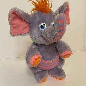 Op de afbeelding: Een pluche olifant in grijstinten, roze en oranje. De olifant heeft grote oren met roze binnenkanten, blauwe ogen en een toefje oranje haar. Het heeft een gestreepte roze en paarse band rond zijn middel.