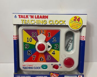 Könnte beinhalten: Ein weißes Talk 'n Learn Teaching Clock Spielzeug in der Originalverpackung. Das Zifferblatt ist mehrfarbig mit den Zahlen 1-12. Es hat einen roten Knopf und einen Griff. Die Verpackung enthält den Text "24 lustige Aktivitäten mit authentischer Stimme und Melodien."