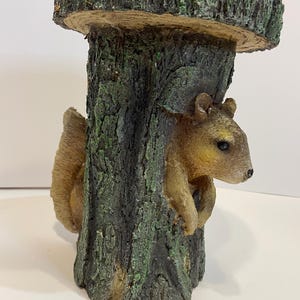 May include: A detailed squirrel ornament emerging from a tree stump. The tree stump is painted in shades of green and brown, with a textured bark effect. The squirrel has a bushy tail and is looking to the side. The ornament is designed to look like a small table.