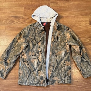 May include: A long-sleeve jacket featuring a camouflage pattern of brown, tan, and green leaves and branches. The jacket includes a light gray jersey hood, a dark brown inner lining, a button-up front closure, and two chest pockets with flaps.