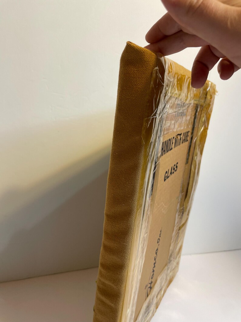 May include: A wrapped canvas art piece with a mustard-coloured fabric edge. The cardboard packaging is sealed with clear tape and features the text "HANDLE WITH CARE" and "GLASS". The art piece is from "Homeco, Inc."