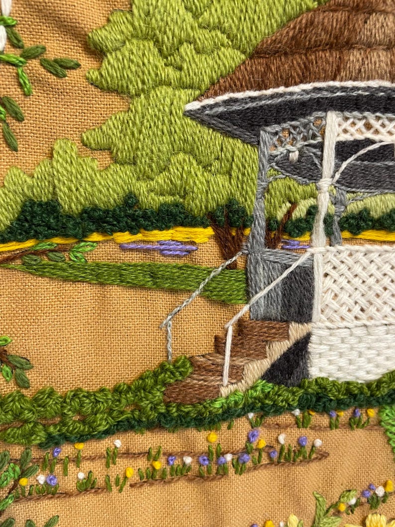 May include: Embroidered landscape scene showing a gazebo with a brown roof, grey supports, and white lattice. The scene includes green trees, a small pond, and a path with flowers. The embroidery is on a tan fabric.