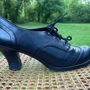 Könnte beinhalten: Schwarzer Lederschuh mit Absatz und Schnürdesign. Der Schuh hat eine runde Spitze, dekorative Nähte und einen Blockabsatz. Der Schuh steht auf einer gewebten Oberfläche mit grünem Hintergrund.