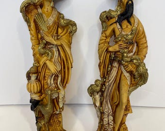 May include: Two antique Asian figurines. The female figure wears a yellow robe, black hat, and holds a fan. The male figure wears a hat and holds a scroll. Both figures have gold accents and are on ornate bases.