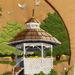 May include: Embroidered artwork showing a gazebo in a park. The gazebo has a brown and white chequered roof, white latticework, and grey pillars. Green trees and blue and yellow flowers surround the gazebo. Three grey birds are flying in the sky.