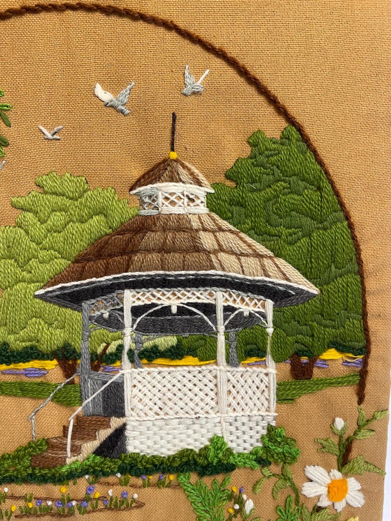 May include: Embroidered artwork showing a gazebo in a park. The gazebo has a brown and white chequered roof, white latticework, and grey pillars. Green trees and blue and yellow flowers surround the gazebo. Three grey birds are flying in the sky.