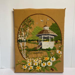 May include: Embroidered wall hanging depicting a detailed landscape scene. A white gazebo with a brown roof is surrounded by green trees and a field of daisies. The artwork is set within a brown circular frame on a tan background.