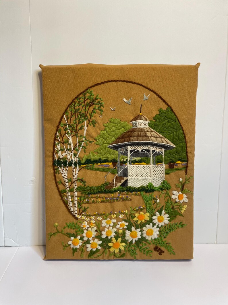 May include: Embroidered wall hanging depicting a detailed landscape scene. A white gazebo with a brown roof is surrounded by green trees and a field of daisies. The artwork is set within a brown circular frame on a tan background.