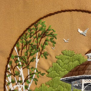 May include: Embroidered landscape scene with a birch tree, gazebo, and birds, stitched on tan fabric. The birch tree has white bark and green leaves. The gazebo is grey and white. The scene is framed by a brown stitched border. The embroidery uses green, white, and brown thread.