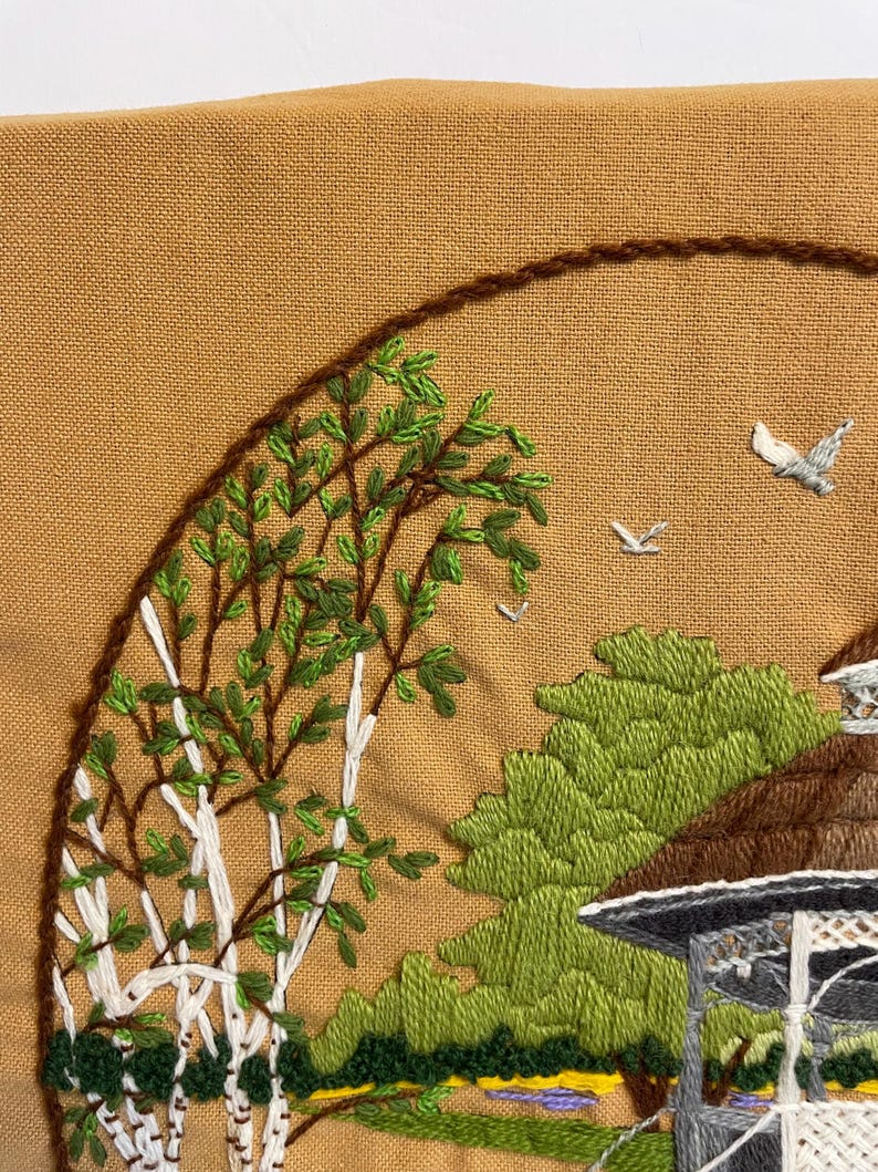 May include: Embroidered landscape scene with a birch tree, gazebo, and birds, stitched on tan fabric. The birch tree has white bark and green leaves. The gazebo is grey and white. The scene is framed by a brown stitched border. The embroidery uses green, white, and brown thread.