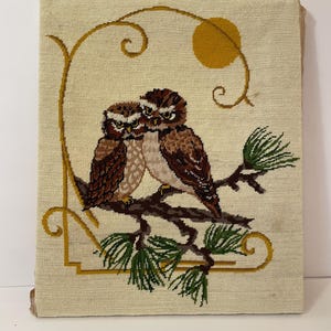 May include: A vintage needlepoint wall hanging featuring two brown owls perched on a branch with green needles. The artwork includes a yellow sun and a gold-colored decorative border on a cream-colored background. The piece is rectangular.