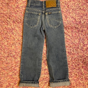 Pode incluir: Jeans azuis de ganga com o logótipo Lee no bolso traseiro e um remendo de couro castanho. Os jeans têm um design clássico de cinco bolsos e são dobrados nos tornozelos. Os jeans são provavelmente para uma criança.