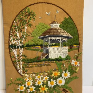 May include: Embroidered wall hanging depicting a garden scene. A white gazebo with a brown roof is surrounded by green trees, a birch tree, and daisies. The artwork is set within a brown circular frame, creating a framed effect.