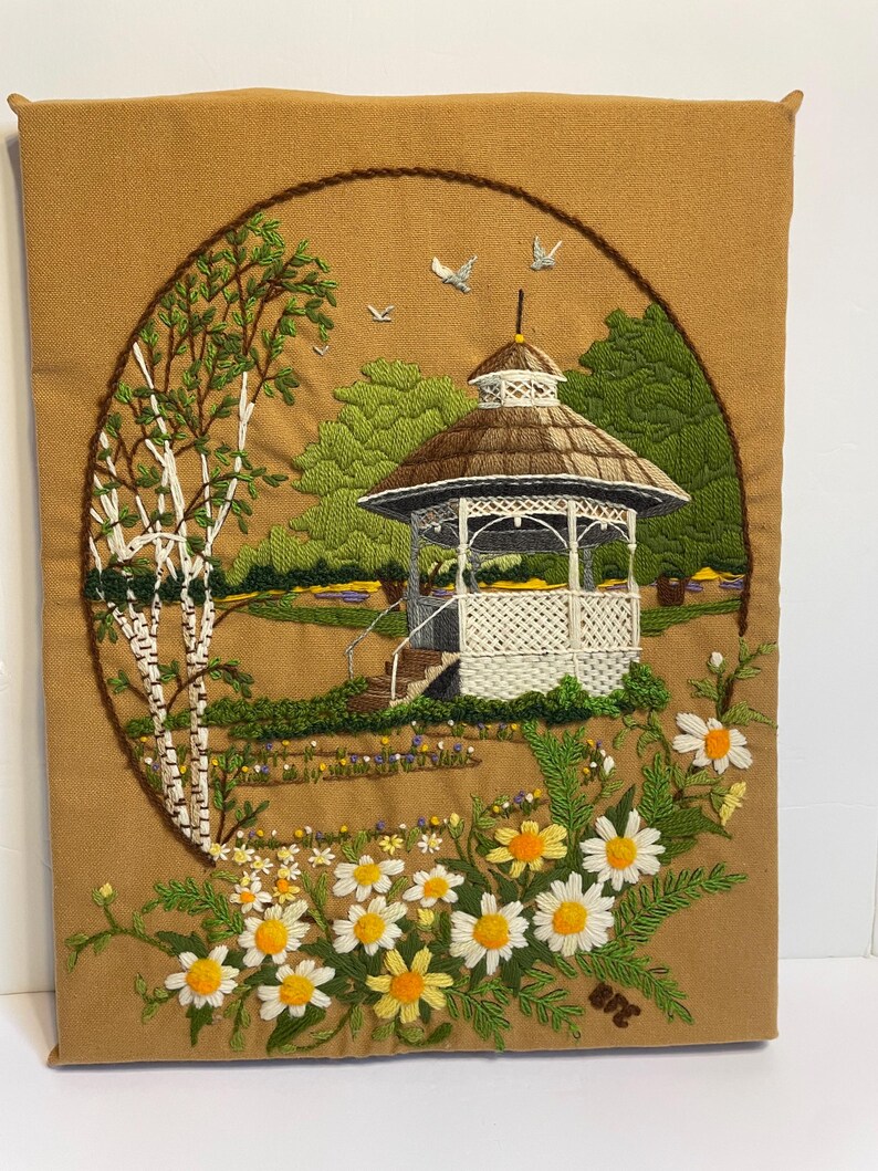 May include: Embroidered wall hanging depicting a garden scene. A white gazebo with a brown roof is surrounded by green trees, a birch tree, and daisies. The artwork is set within a brown circular frame, creating a framed effect.
