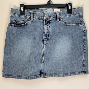 May include: A light blue denim skirt with a classic five-pocket design. The skirt has a button and zip closure, belt loops, and a faded wash. The skirt is hanging on a black hanger.
