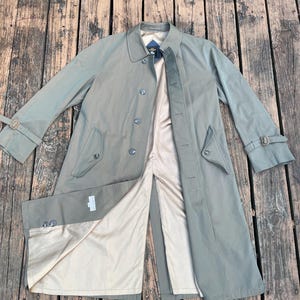 May include: A long, green trench coat with a brown belt and a beige lining. The coat has a double-breasted closure and a single button on each cuff.