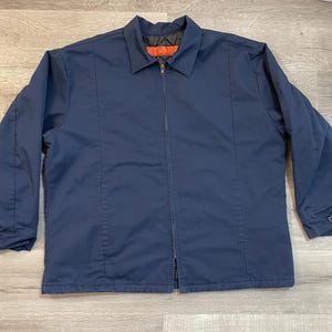 May include: A navy blue work jacket with a full-length zipper closure and a collared neckline. The jacket has two vertical seams on the front and buttoned cuffs. The interior collar tag is orange. This jacket is suitable for work or casual wear.
