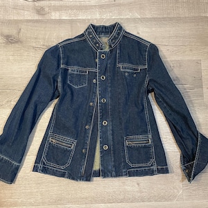 May include: A dark blue denim jacket with a button-up front, two chest pockets, and two lower pockets with zippers. The jacket has long sleeves with buttoned cuffs and a collarless neckline.