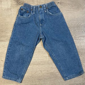 May include: Blue denim toddler jeans with an elastic waistband and classic five-pocket design. The jeans have a button and zipper closure, and are made of a durable denim material. The pants are designed for comfort and everyday wear.