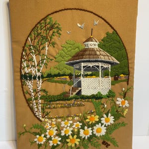 May include: Embroidered wall hanging depicting a scenic landscape. A white gazebo with a brown roof is surrounded by green trees and a field of daisies. The artwork is framed by a brown border and set against a tan background.