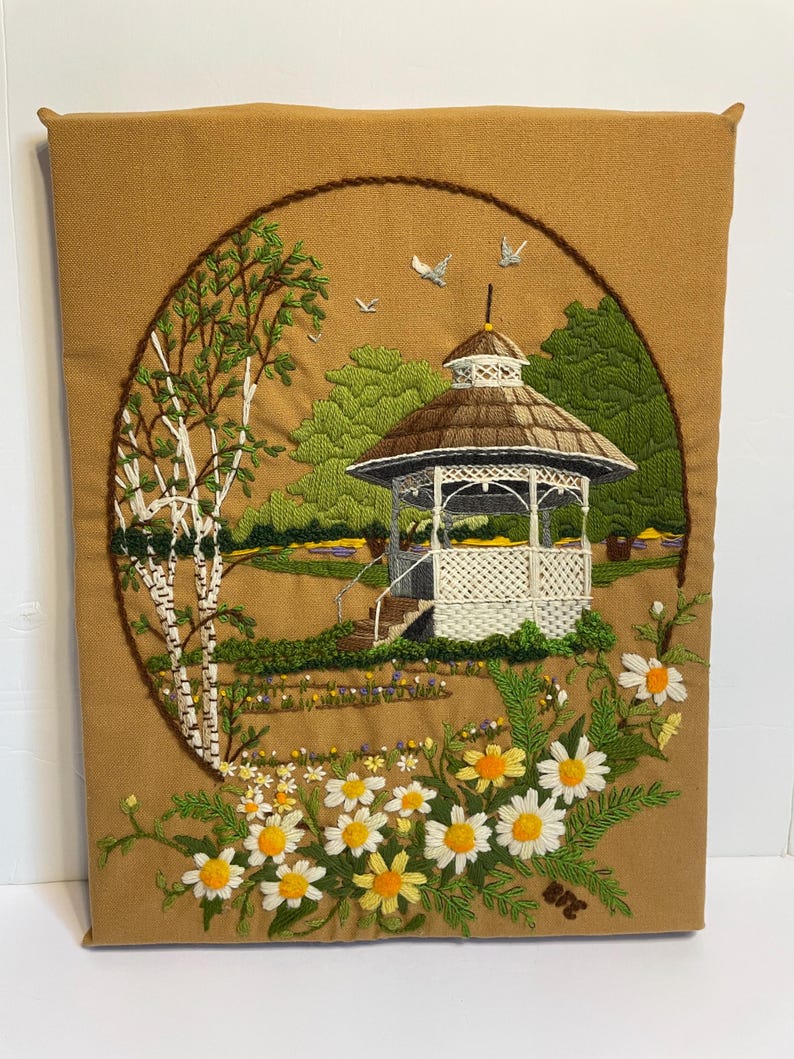 May include: Embroidered wall hanging depicting a scenic landscape. A white gazebo with a brown roof is surrounded by green trees and a field of daisies. The artwork is framed by a brown border and set against a tan background.