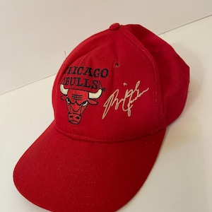 Puede incluir: Gorra de béisbol roja con el logotipo de los Chicago Bulls y una firma blanca. La gorra tiene una visera curva y un botón en la parte superior. El logotipo de los Chicago Bulls es una cabeza de toro roja con las palabras "CHICAGO BULLS" encima.