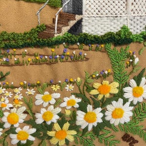 May include: Embroidered textile art depicting a garden scene. The artwork features a white picket fence, stone steps, and a variety of embroidered flowers, including daisies with yellow centres and white petals, set against a tan background.