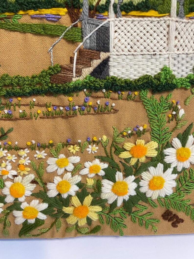 May include: Embroidered textile art depicting a garden scene. The artwork features a white picket fence, stone steps, and a variety of embroidered flowers, including daisies with yellow centres and white petals, set against a tan background.