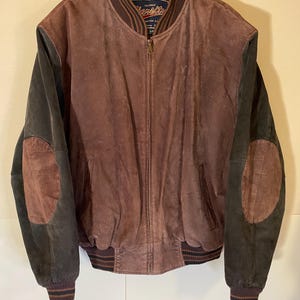 May include: A vintage brown leather jacket with dark green sleeves and elbow patches. The jacket has a ribbed collar, cuffs, and hem, and a zipper closure. The word "Varsity" is visible on the collar.