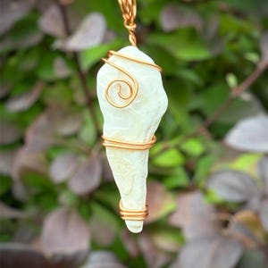 May include: A pendant featuring a cream-colored, organically shaped stone wrapped with copper wire. The wire forms a spiral design at the top and wraps around the stone's body. The pendant hangs from a black cord.