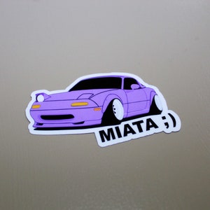 May include: A purple sticker of a cartoon car with the text "MIATA :)" underneath.