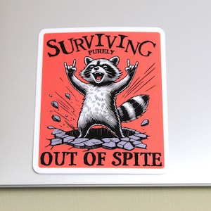 K&ouml;nnte beinhalten: Rechteckiger Aufkleber mit einer Cartoon-Waschb&auml;r-Illustration. Der Waschb&auml;r bricht durch eine Oberfl&auml;che, die Arme in einer Rock'n'Roll-Geste erhoben. Der Aufkleber hat den Text "SURVIVING PURELY OUT OF SPITE" in Schwarz und Wei&szlig;.