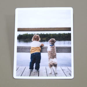 May include: A young child and a small dog stand side-by-side on a wooden dock, looking out at a lake. The child is wearing a yellow and white shirt, blue jeans, and brown shoes. The dog is brown and white with a blue collar.