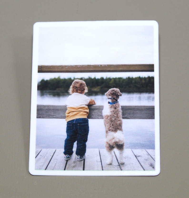 May include: A young child and a small dog stand side-by-side on a wooden dock, looking out at a lake. The child is wearing a yellow and white shirt, blue jeans, and brown shoes. The dog is brown and white with a blue collar.