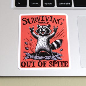 K&ouml;nnte beinhalten: Ein rechteckiger Aufkleber mit rotem Hintergrund und einer Waschb&auml;r-Illustration. Der Waschb&auml;r bricht durch eine Oberfl&auml;che, die Arme in einer Rock'n'Roll-Geste erhoben. Der Text "SURVIVING PURELY OUT OF SPITE" ist in Schwarz gedruckt.