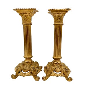 May include: Two antique gold-colored candle holders. Each candle holder has a fluted column design, a decorative base, and a detailed top. The candle holders are made of metal and are displayed against a white background.