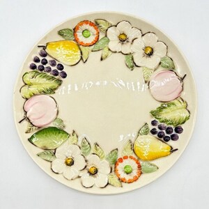 May include: A white ceramic plate with a raised design of fruit and flowers. The plate features pears, apples, grapes, and flowers in shades of pink, yellow, green, and orange.