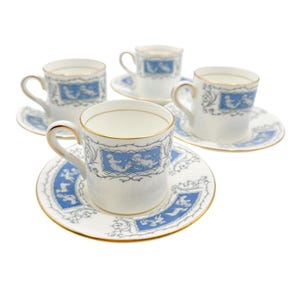 May include: Four white teacups and saucers with a blue and gray design. The cups and saucers have a gold trim. The cups have a handle and a blue rectangular design with white figures. The saucers have a blue and gray border.
