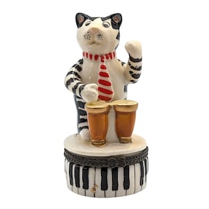 May include: A black and white cat figurine playing the bongos. The cat is wearing a red and white striped tie. The figurine is sitting on a base that looks like a piano keyboard.