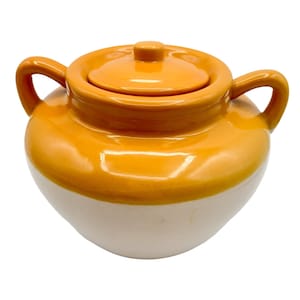 May include: A ceramic pot with a lid, featuring a two-tone design. The top half is a vibrant orange, while the bottom half is a creamy white. The pot has two handles on the sides and a rounded shape.