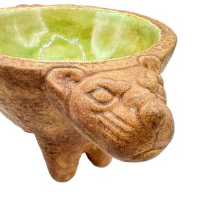 May include: A ceramic bowl with a green glaze inside and a brown, textured exterior. The bowl is shaped like an animal's head with a snout and ears.