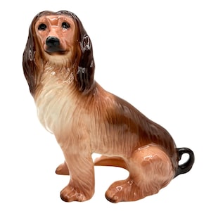 May include: A ceramic figurine of a brown and white Afghan Hound sitting with its head turned to the side. The figurine is detailed with realistic fur and features.