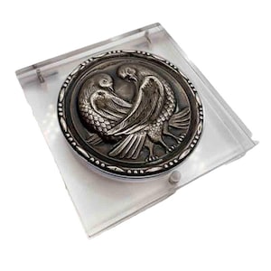 May include: A silver-toned round compact featuring a detailed relief of two doves facing each other. The compact is encased in a clear plastic sleeve. The doves are intricately designed with detailed feathers and a decorative border.