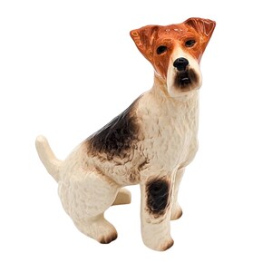 May include: A ceramic figurine of a wirehaired fox terrier dog sitting with its head tilted down. The dog is white with brown and black markings.