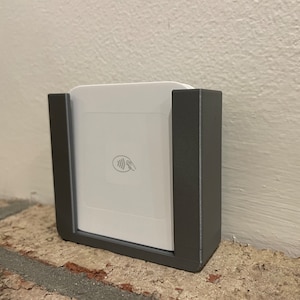 May include: A gray plastic wall mount for a white square-shaped credit card reader with a contactless payment symbol.